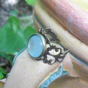 Puede incluir: Un anillo con una piedra cabuj&oacute;n azul claro engastada en un dise&ntilde;o de filigrana de bronce.