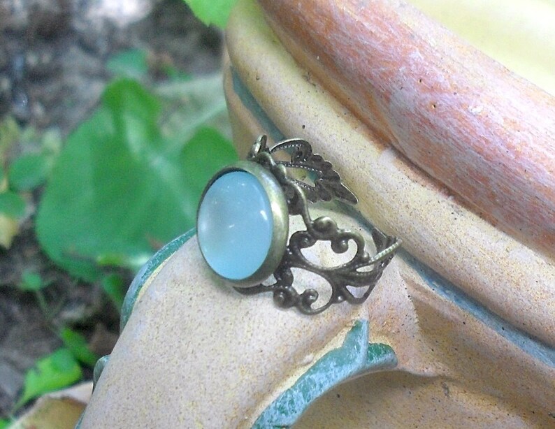 Puede incluir: Un anillo con una piedra cabuj&oacute;n azul claro engastada en un dise&ntilde;o de filigrana de bronce.