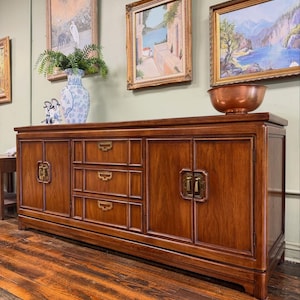 May include: A long wooden sideboard featuring a rich brown finish, two side cabinets with double doors, and three central stacked drawers. The piece is accented with rectangular brass hardware pulls and decorative trim on the door and drawer fronts.
