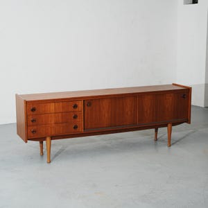 May include: A long, rectangular wooden sideboard featuring three stacked drawers on the left side with circular wooden pulls. The right side includes two sliding cabinet doors with matching circular pulls. The unit is supported by four tapered wooden legs.