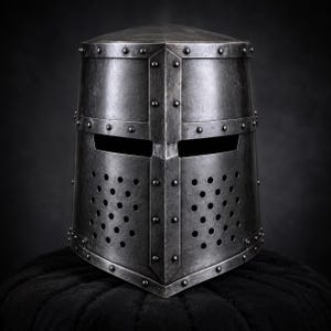 Puede incluir: Un yelmo de metal gris oscuro con una parte superior plana, una barra de refuerzo vertical central y dos ranuras horizontales para los ojos. La sección inferior del casco incluye dos paneles con orificios de ventilación circulares y remaches.