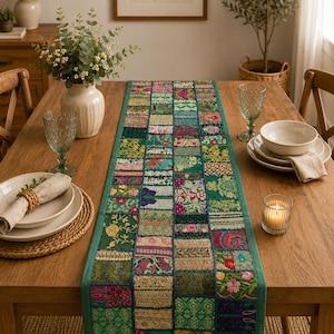 May include: A rectangular table runner featuring a patchwork design of various green, pink, and gold fabric squares. The runner is composed of multiple embroidered and patterned textile pieces stitched together with a solid green border trim.