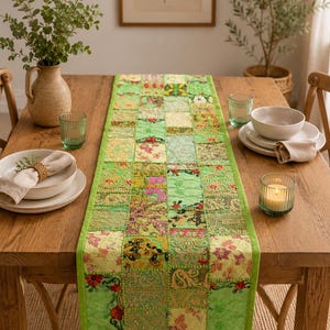 May include: A rectangular table runner featuring a patchwork design of various green fabric squares. Each square displays intricate embroidery, floral patterns, and metallic thread detailing. The runner is bordered by a solid light green fabric trim.