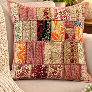 May include: A square throw cushion featuring a patchwork design of various embroidered fabrics in shades of pink, gold, orange, and green. The surface is composed of rectangular panels with intricate floral patterns, beadwork, and metallic thread accents.