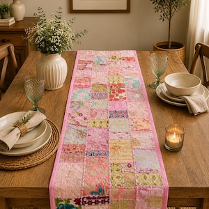 May include: A rectangular table runner featuring a patchwork design of various pink, floral, and embroidered fabric squares. The runner is bordered by a solid pink fabric trim and showcases intricate stitching connecting each individual textile piece.