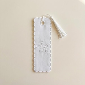 May include: A white rectangular bookmark with scalloped edges, featuring an embroidered monogram of the letters AR in the center. The bookmark includes a circular hole at the top with an attached white fabric tassel made of thin thread.