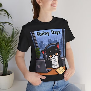 以下が含まれることがあります： ヘッドホンをつけた黒猫が雨の日に窓辺でノートパソコンに向かっているグラフィックがプリントされた黒の半袖Tシャツ。「HOT COFFEE BETTER DAYS」と書かれたマグカップを猫が持ち、黄色い付箋には「BIG PLANS 0」と記されている。