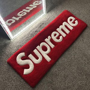 May include: A rectangular red rug featuring the word Supreme in large, raised white letters. The rug has a textured, tufted surface and is positioned near a mirror with a light-up frame, which reflects the red and white text of the rug.