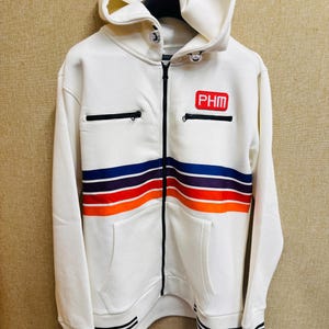 May include: A white zip-up hoodie featuring a red rectangular patch with the text PHM on the chest. The garment includes two horizontal zippered pockets, a multi-colored stripe pattern in navy, purple, and orange, and black striped cuffs and hem.