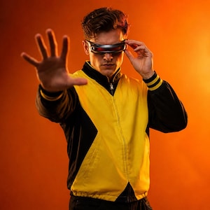 Puede incluir: Una chaqueta con cremallera que presenta un panel frontal amarillo brillante contrastado por mangas y paneles laterales negros, combinada con una visera negra elegante que cubre los ojos. La chaqueta incluye cuello alto y puños acanalados.