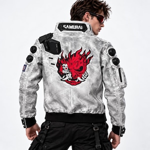 Puede incluir: Una chaqueta de cuero gris claro con aspecto desgastado que presenta un gran gráfico rojo y negro de una máscara de demonio estilizada en la espalda. El cuello muestra el texto SAMURAI en letras blancas. Incluye detalles negros en los hombros.