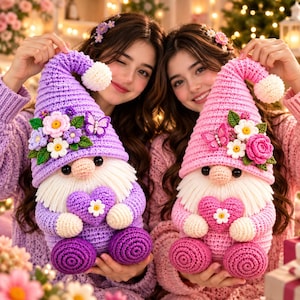 Puede incluir: Dos gnomos de ganchillo hechos a mano con texturas de hilo suave. El gnomo izquierdo es morado con barba blanca, un corazón y detalles de flores y mariposas. El gnomo derecho es rosa con barba blanca, un corazón y detalles de flores.