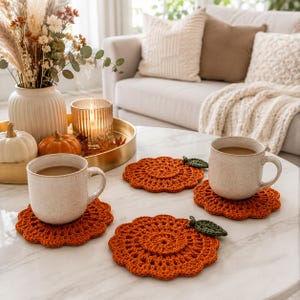 May include: A set of round, orange, crocheted coasters shaped like pumpkins, each featuring a small, dark green crocheted leaf attached to the edge. The coasters are displayed on a white surface, with two of them holding ceramic mugs filled with coffee.