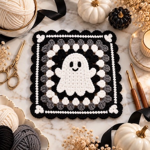 Può includere: Un quadrato all'uncinetto che presenta un fantasma bianco centrale con occhi e sorriso neri. Il design è circondato da strati concentrici di punti di lana nera, bianca e grigia, rifinito con un bordo smerlato nero e cuori bianchi.