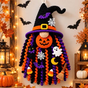 May include: A crocheted wall hanging featuring a gnome wearing a black witch hat with an orange band and yellow buckle. The gnome has a round beige nose, a bright orange jack-o'-lantern face, and long curly black, orange, and purple yarn hair.