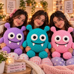 May include: Three plush monster toys made of crocheted yarn. The left toy is purple, the center toy is teal, and the right toy is pink. Each monster features two small horns, large white circular eyes with black pupils, and two white fangs.
