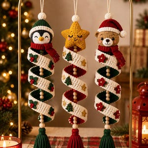 Op de afbeelding: Drie hangende gehaakte ornamenten aan een gouden standaard. Links een pinguïn met een groene muts, in het midden een gele ster, rechts een beer met een rode muts. Elk heeft een wit spiraallint met feestelijke motieven en een kwastje.