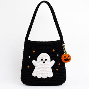 Pode incluir: Uma bolsa de ombro de crochê preta com uma aplicação de fantasma branco no centro, acentuada com pequenas estrelas bordadas em laranja e branco. Um pequeno pingente de abóbora de crochê laranja está pendurado em um fecho de metal na alça.