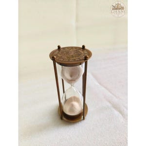 May include: An antique-style hourglass with a metallic bronze frame and clear glass bulbs containing light-colored sand. The top circular plate features an engraved clock face design with Roman numerals. The logo GHANI CRAFT is visible.