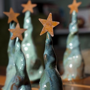 May include: Five ceramic Christmas trees with a rustic, glazed finish. Each tree is topped with a star-shaped ornament made of rusty metal.