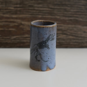 May include: A handmade, cylindrical ceramic vase in a speckled blue glaze. A black dinosaur skeleton is etched on the side. The rim and base have a brown, unglazed finish. The vase is approximately 4 inches tall.