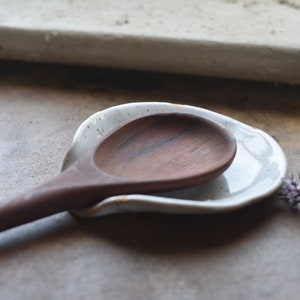 May include: A white ceramic spoon rest with a wooden spoon resting inside. The spoon rest has a speckled glaze and the spoon is a dark brown color.