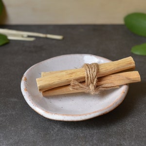 May include: Three bundles of palo santo sticks tied together with twine, resting on a white ceramic dish. The dish has a speckled glaze and a slightly irregular shape.