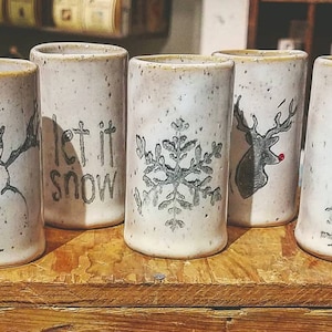 May include: Five white ceramic mugs with winter-themed designs. The mugs feature a snowman, a snowflake, a reindeer, a pine tree, and the words "let it snow".