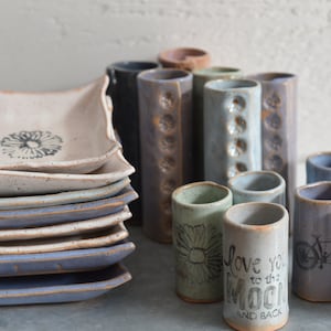 May include: A stack of eight white ceramic square dishes with blue trim and a black floral design.  A collection of 12 ceramic cylinder vases in various shades of blue, green, and brown with different designs.  Some of the vases have a floral design, a bicycle design, and a cat design.  One vase has the text "Love you to the moon and back".