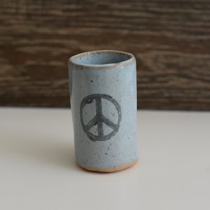 May include: A light blue ceramic shot glass with a peace sign design. The glass has a speckled glaze.