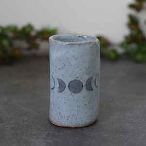 May include: A light blue ceramic vase with a speckled glaze and a design of five crescent moons in black.