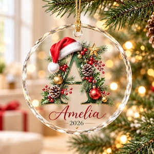 May include: A circular clear glass Christmas ornament featuring a large letter A decorated with pine branches, red berries, pinecones, gold ornaments, and a Santa hat. The name Amelia and the year 2026 are printed in red script and black text.