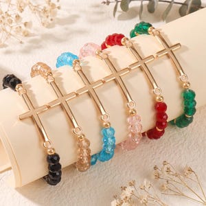 May include: A collection of six stretch bracelets featuring faceted crystal beads in black, beige, blue, pink, red, and green. Each bracelet includes a central gold-tone metal cross charm connected by small gold-tone metal beads and jump rings.