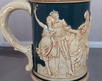 Op de afbeelding: Een keramische bierpul met een crèmekleurige handgreep en deksel. De donkerblauwe body heeft een reliëf van figuren in historische kleding. De basis en het deksel hebben decoratieve bloempatronen.
