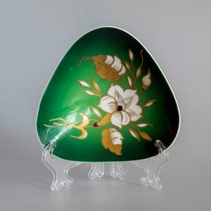 Op de afbeelding: Een driehoekige kom met een diepgroene afwerking, voorzien van een centraal wit bloemmotief met goudkleurige bladaccenten en stelen. De kom wordt gepresenteerd op een doorzichtige, driepotige kunststof standaard, wat het gladde oppervlak benadrukt.