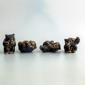 Pode incluir: Quatro pequenas figuras de urso em cerâmica dispostas em fila. Cada urso tem um acabamento em esmalte preto fosco com reflexos castanho-dourados que acentuam a textura do pelo e os traços faciais. Os ursos estão em várias posições.