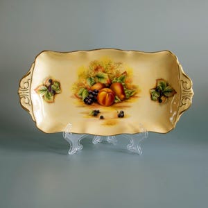 May include: A rectangular porcelain serving tray with a cream-coloured base and scalloped edges featuring gold trim. The centre displays a painted arrangement of peaches, grapes, and green leaves, with smaller fruit clusters on the side handles.