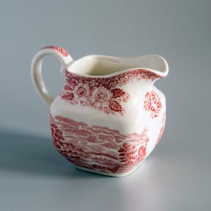 Pode incluir: Uma leiteira de cerâmica cor de creme com um padrão de transferência vermelho. O design inclui uma cena de paisagem detalhada com árvores e colinas no corpo, complementada por uma borda floral e uma alça com detalhes em vermelho.