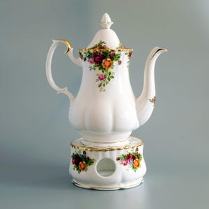 May include: A white porcelain teapot with a matching warmer stand, featuring a scalloped base and body. The set is decorated with a colourful floral pattern of red, yellow, and pink roses with green leaves, accented by gold trim on the edges and handle.