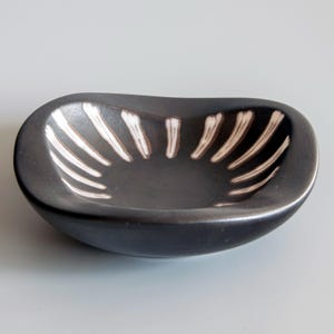Può includere: Una piccola ciotola in ceramica di forma ovale e poco profonda, con esterno ed interno nero opaco. Il bordo interno presenta una serie di brevi tratti di pennello bianchi e verticali che si irradiano verso il centro della ciotola.