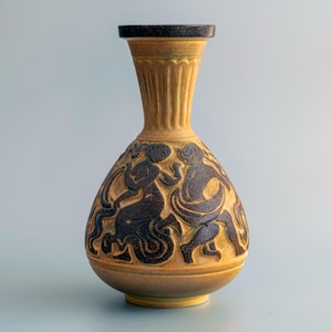 Pode incluir: Um vaso de cerâmica com esmalte amarelo mostarda e borda marrom escura. O corpo apresenta um design em relevo de figuras marrom escuras em movimento sobre o fundo amarelo. O pescoço do vaso tem detalhes estriados verticais.