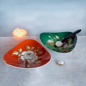 May include: Two triangular ceramic dishes, one orange and one green, each featuring a white and gold floral pattern. The orange dish holds a gold ring, while the green dish contains a wristwatch with a dark strap and a gold chain.