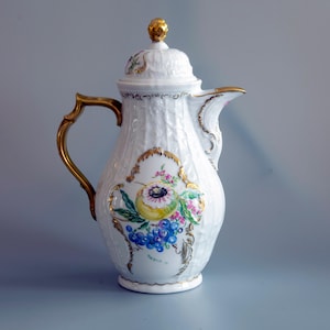 Może przedstawiać: Biały dzbanek do kawy z porcelany o fakturze plecionki, ze złotym uchwytem i złotym zwieńczeniem na pokrywce. Korpus ozdobiony jest ręcznie malowanym motywem owocowym, przedstawiającym winogrona i żółty owoc, w złotej ramce.