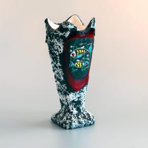May include: A tall, textured vase with a rough, black and white cratered surface. The front features a smooth, glossy, shield-shaped inset in deep red, displaying a graphic illustration of two stylized fish with yellow, blue, and white details.