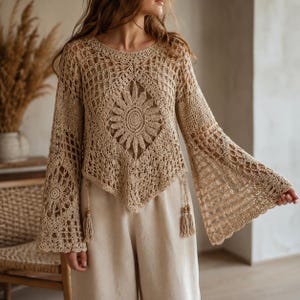 Op de afbeelding: Een beige gehaakte top met lange mouwen, voorzien van een ingewikkeld centraal zonnebloemmotief, een ruitvormige zoom en wijde klokmouwen. Het kledingstuk heeft twee hangende kwastjes met houten kralen bij de taille en een opengewerkt patroon.