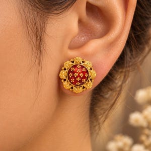 Puede incluir: Un pendiente de botón redondo de oro con un centro circular rojo intenso y un patrón floral dorado en relieve. El borde exterior del pendiente está compuesto por un borde dorado festoneado y texturizado con pequeños detalles de cuentas.