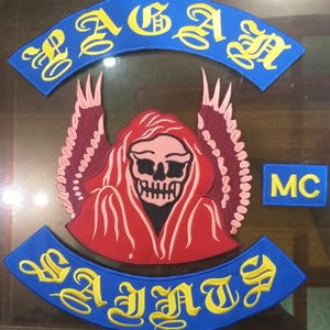 May include: A set of embroidered patches featuring a central red hooded skull figure with wings, flanked by a small blue rectangular patch with yellow MC text, and two curved blue rocker patches with yellow text reading PAGAN and SAINTS.