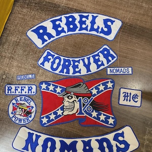 May include: A collection of embroidered patches featuring blue and white text reading REBELS, FOREVER, NOMADS, and R.F.F.R. A central rectangular patch displays a red flag with white stars, a skull wearing a cap, and the text 1%.