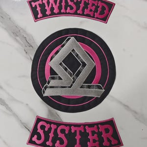 Könnte beinhalten: Ein Set aus drei bestickten Aufnähern, bestehend aus einem schwarzen runden Emblem mit einem silbernen geometrischen Logo und pinken Akzenten. Zwei geschwungene schwarze Aufnäher ergänzen das Emblem, mit dem Wort TWISTED in Pink oben und SISTER in Pink unten.