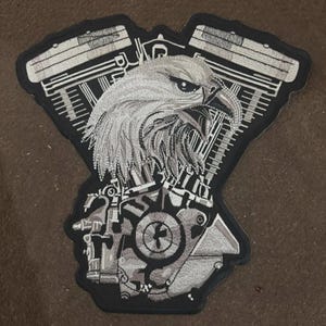 Op de afbeelding: Een grote geborduurde patch met een gedetailleerde zilveren en zwarte adelaarskop, gecentreerd over een gestileerde V-twin motorfietsmotor. Het ontwerp gebruikt ingewikkelde stiksels om de veren, motorcilinders en mechanische onderdelen te definiëren.